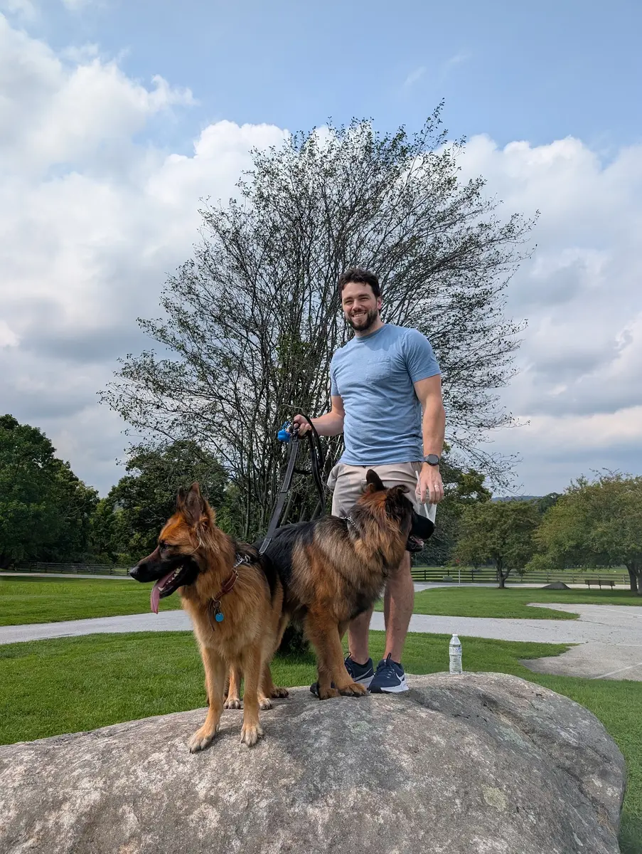 Kyle standing on a big rock in a park with his two long-haired German Shepherds, Bailey and Archie, on leashes beside him