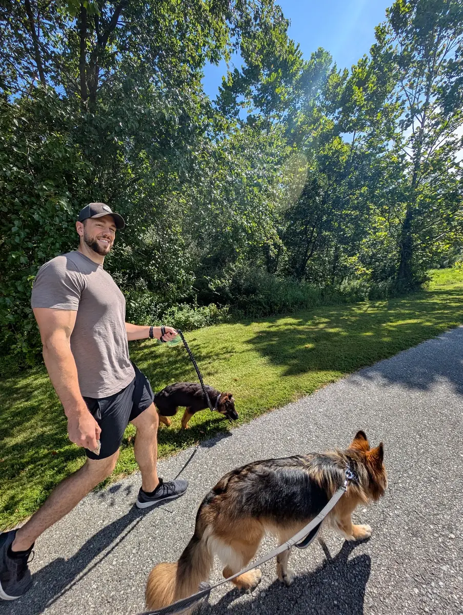 Kyle smiling as he walks Bailey and Archie down a paved, tree-lined trail